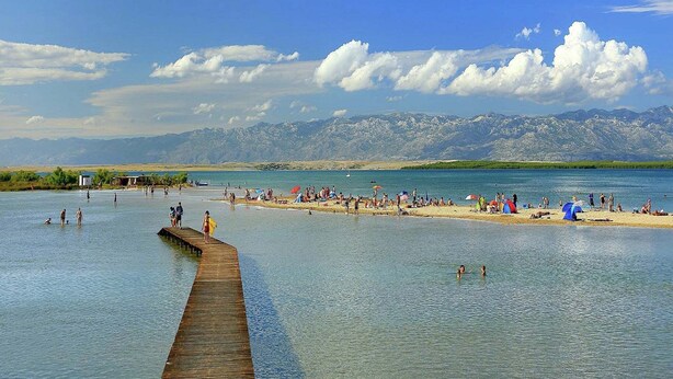 A lakeside beach with a wooden pier extending into the water, surrounded by mountains in the background.