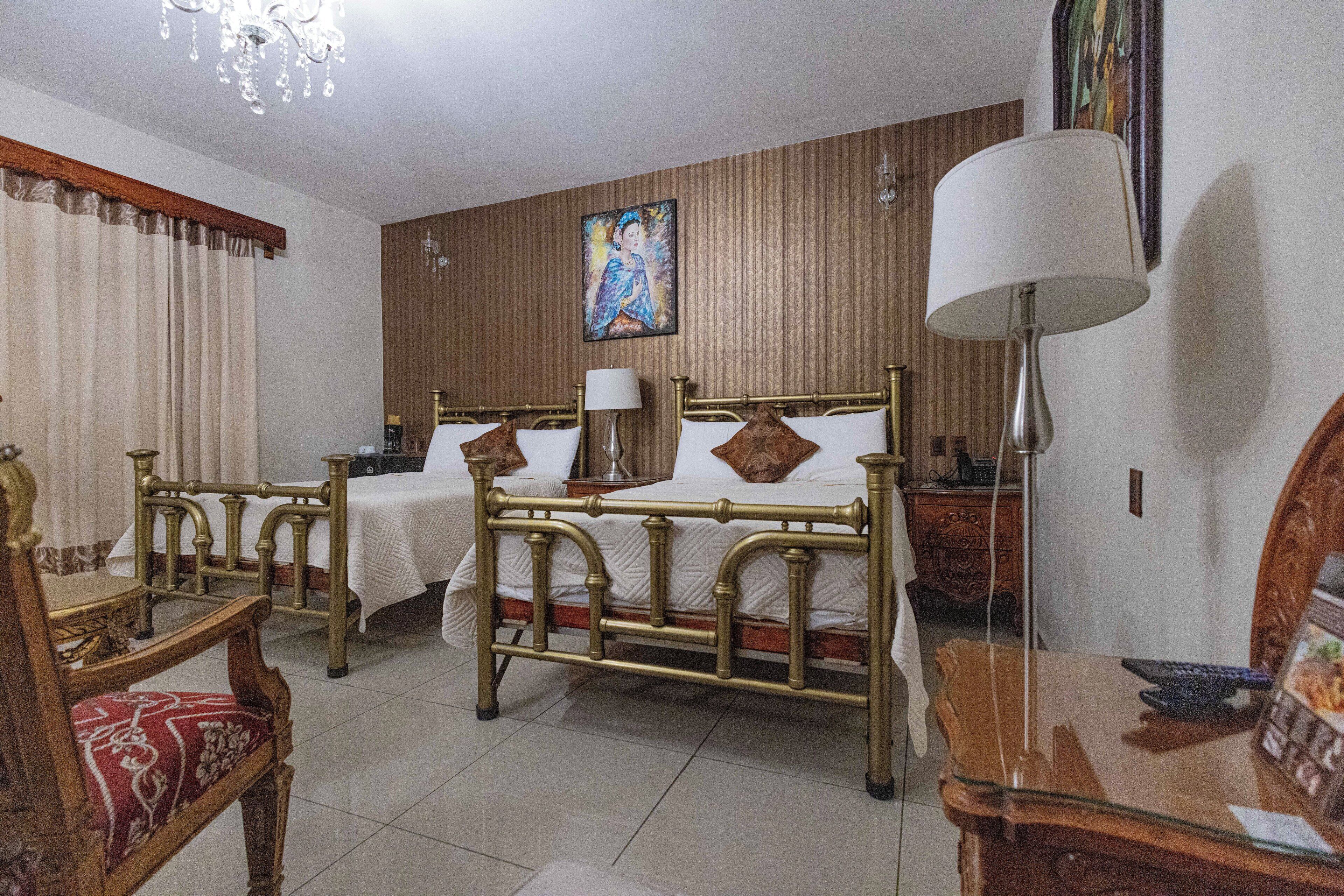 Une chambre d’hôtel avec deux lits, une tête de lit en bois, un lustre, un tableau au mur et un bureau avec une lampe.