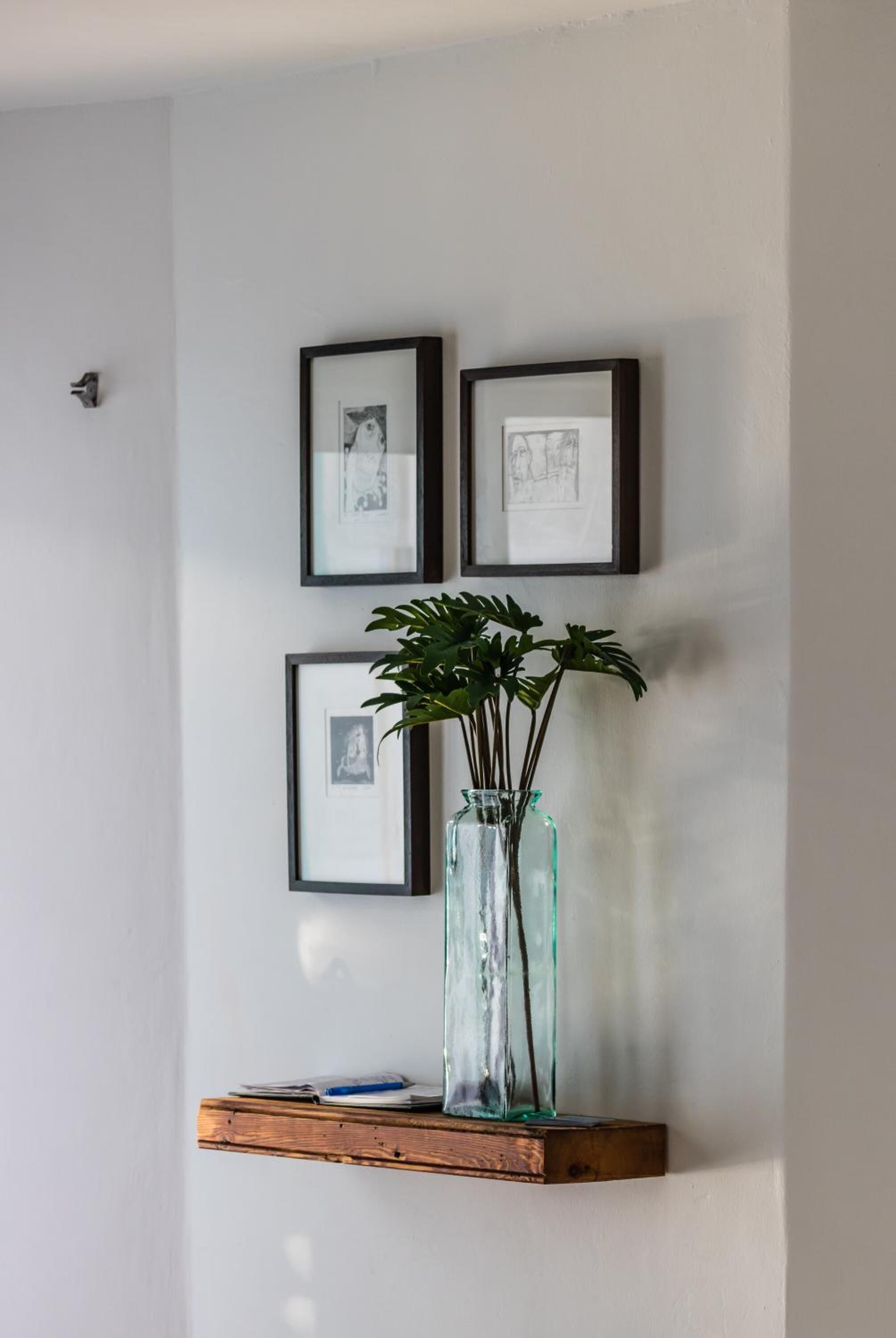 A wall with three framed pictures and a glass vase with plants on a wooden shelf.