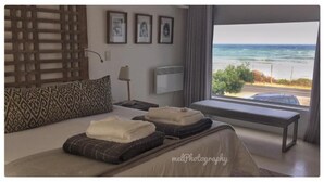 A bedroom with a bed, pillows, a bedside table, a lamp, and a view of the ocean.
