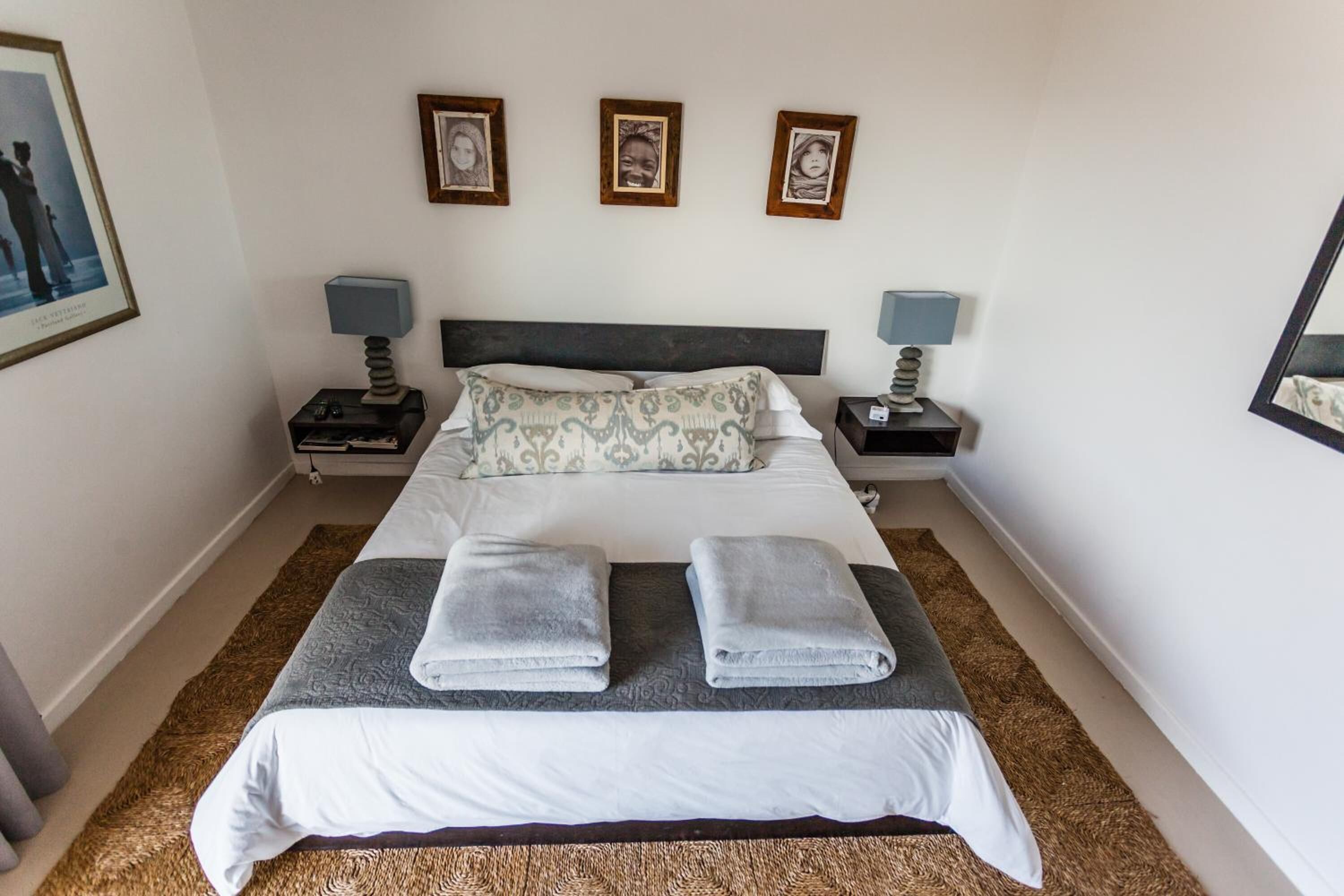 Una cama bien hecha con dos toallas, un cojín estampado y un cabecero. Hay dos mesitas de noche con lámparas y cuadros enmarcados en la pared sobre la cama.