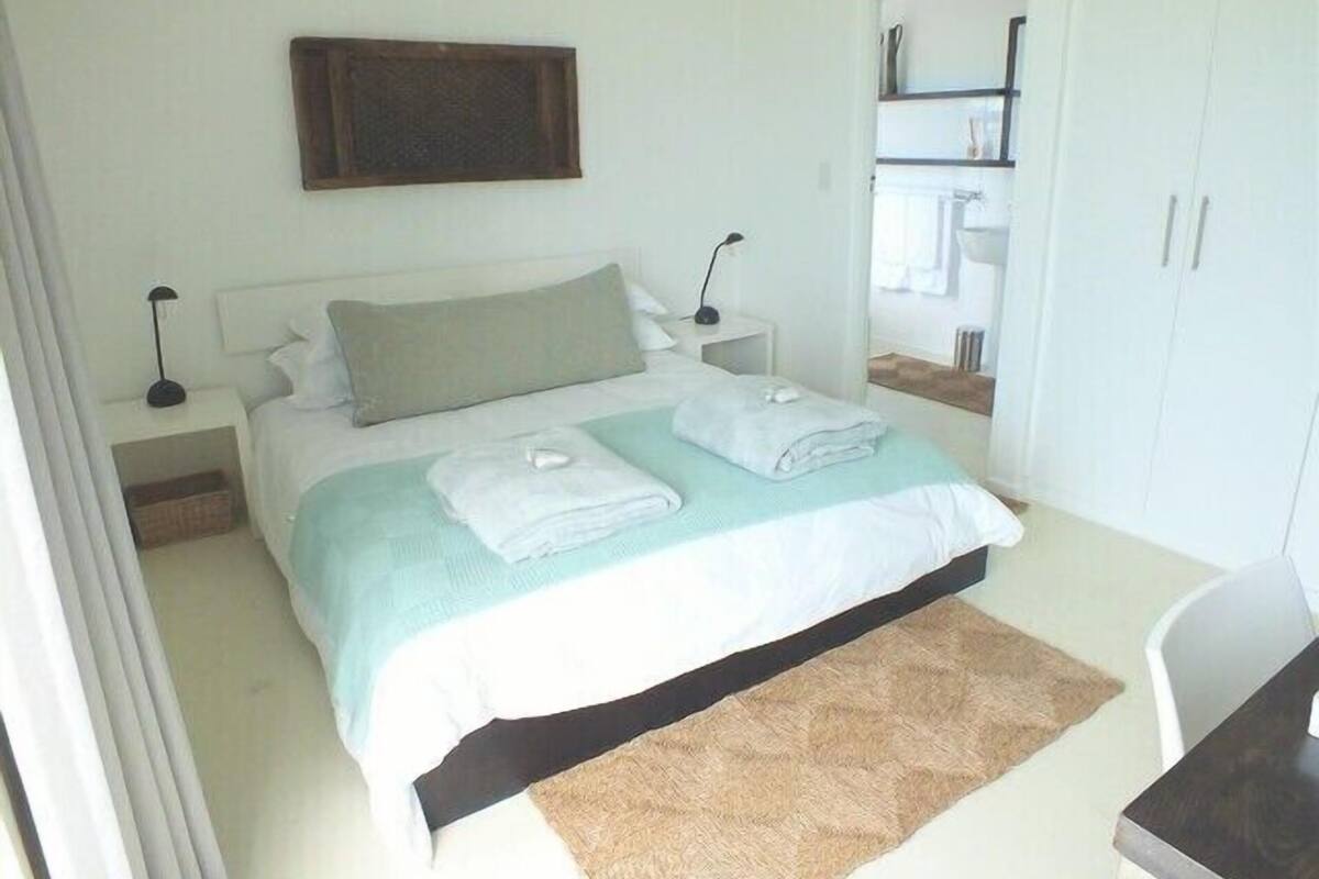 Un lit bien fait, recouvert d’une couvre-lit bleu clair, avec des oreillers blancs et un tapis marron. Une petite table de chevet se trouve à côté du lit, et un miroir est accroché au mur au-dessus du lit.