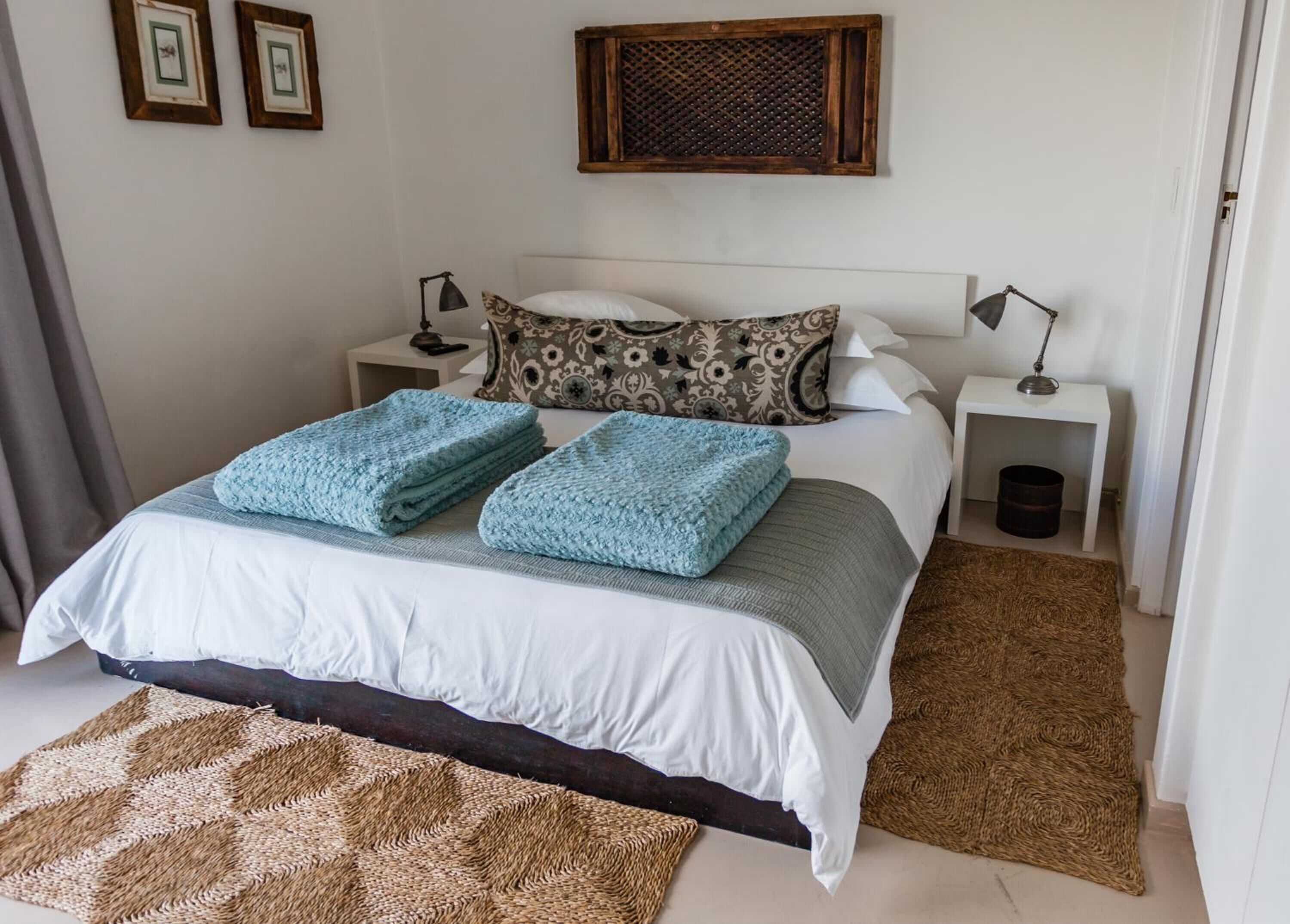 A neatly made bed with a white and grey duvet, two blue folded blankets, and a patterned pillow. There are two bedside tables with lamps, a woven rug on the floor, and a wooden headboard.