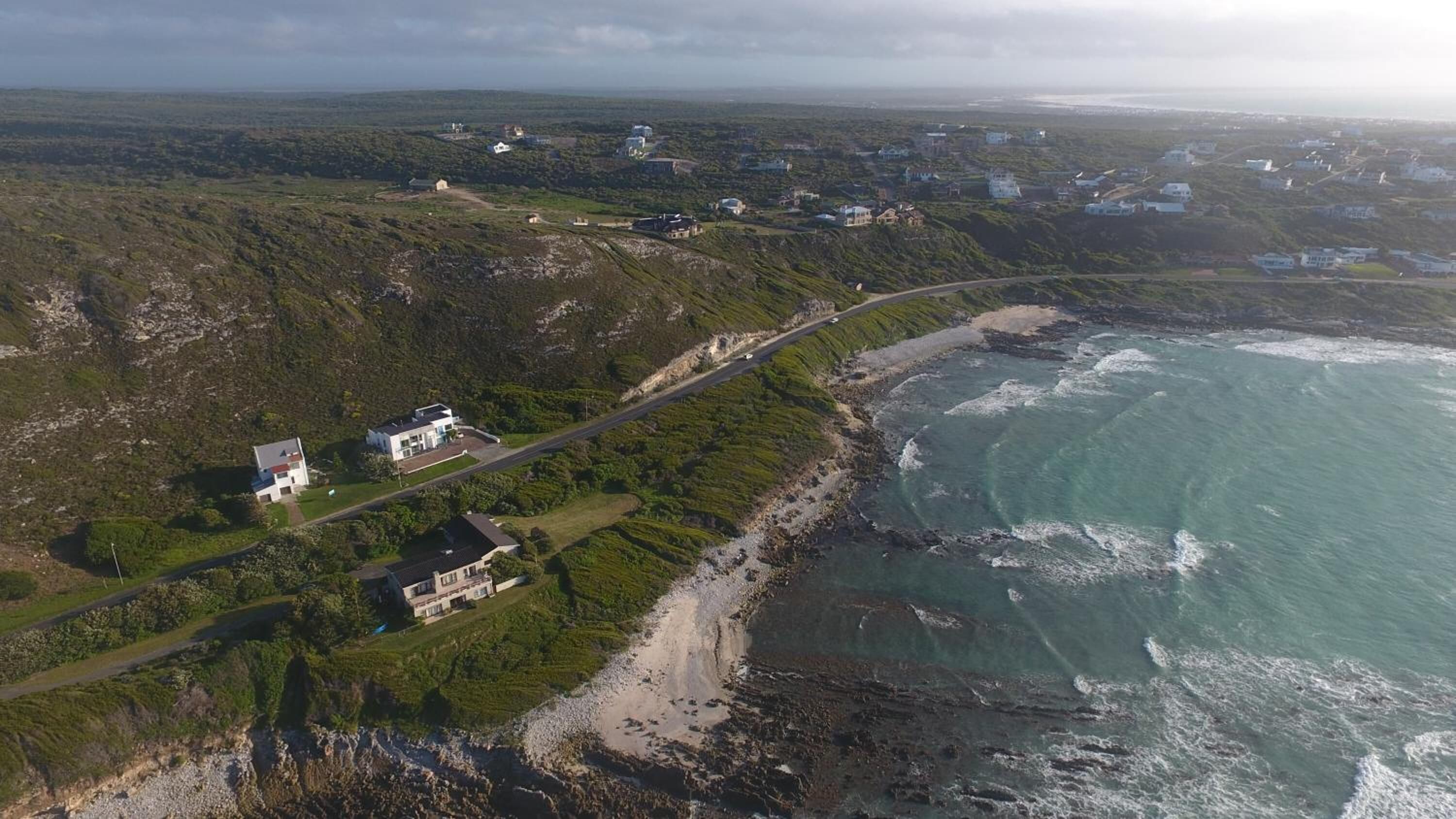 An aerial view of a coastal area with houses, a road, and the ocean.