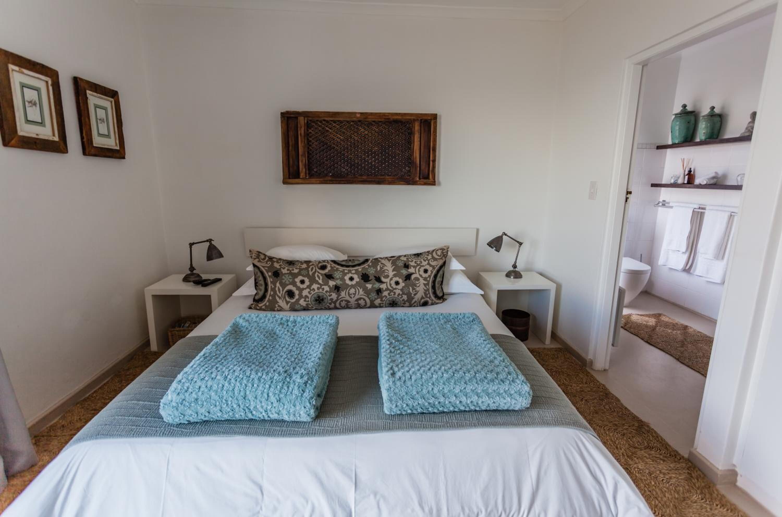 A bedroom with a bed, two bedside tables, a nightstand, a wall-mounted shelf with decorative items, and a door leading to a bathroom.