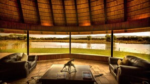 A room with a thatched roof, leather sofas, a wooden coffee table, and a view of a lake and trees. - Mount Etjo Safari Lodge (Omaruru)