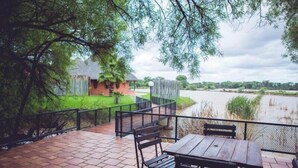 An outdoor patio with a wooden table and chairs, overlooking a river and a fenced area with a small building. - Mount Etjo Safari Lodge (Omaruru)