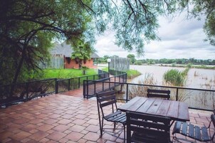 Un patio al aire libre con mesa y sillas de madera, con vista a un río y un área cercada con un pequeño edificio.