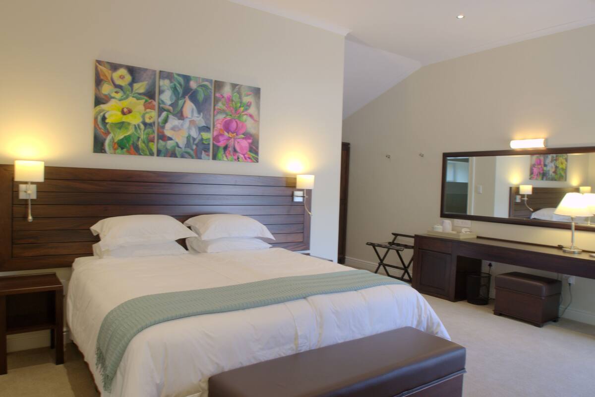 A hotel room with a large bed, a bedside table, a dresser, a mirror, and a painting on the wall.