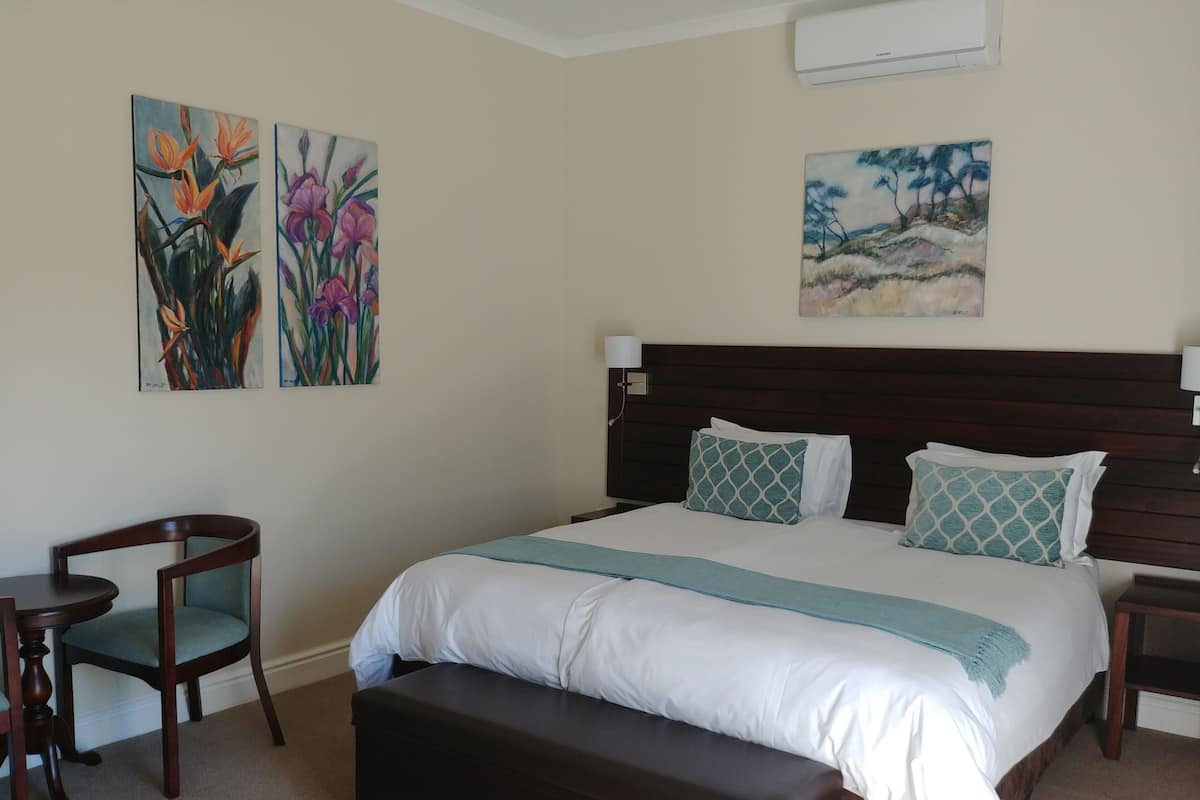 A hotel room with a large bed, a wooden bench, a small round table, and two chairs. There are three paintings on the wall.