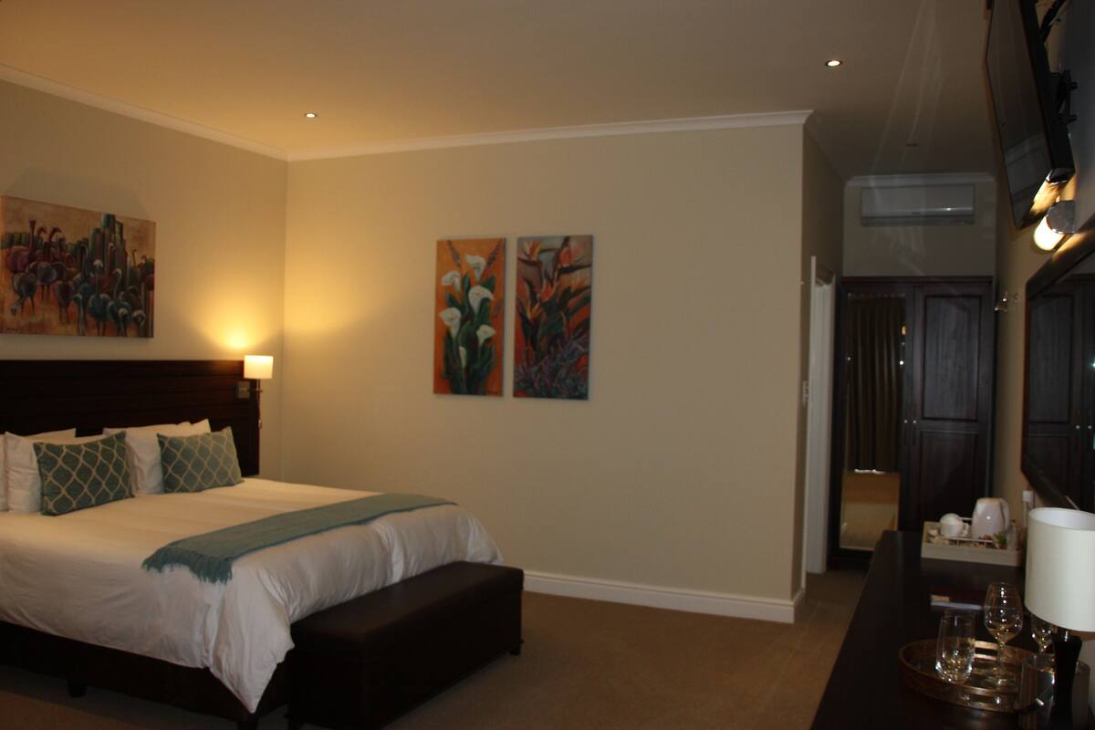 A hotel room with a large bed, a bench, a desk with a lamp, and two paintings on the wall.