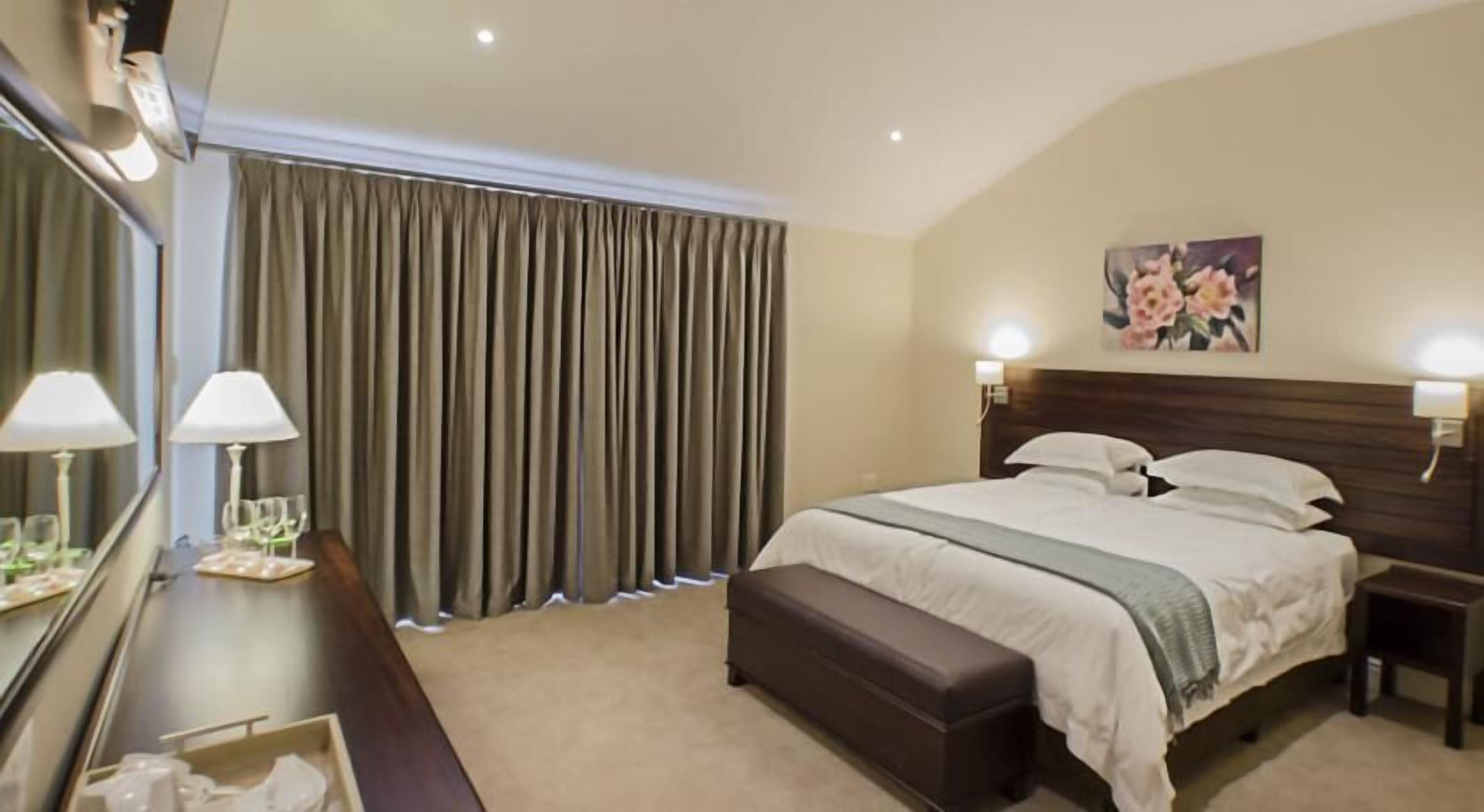 Una habitación de hotel con una cama grande, una mesita de noche, un espejo y una ventana con cortinas.