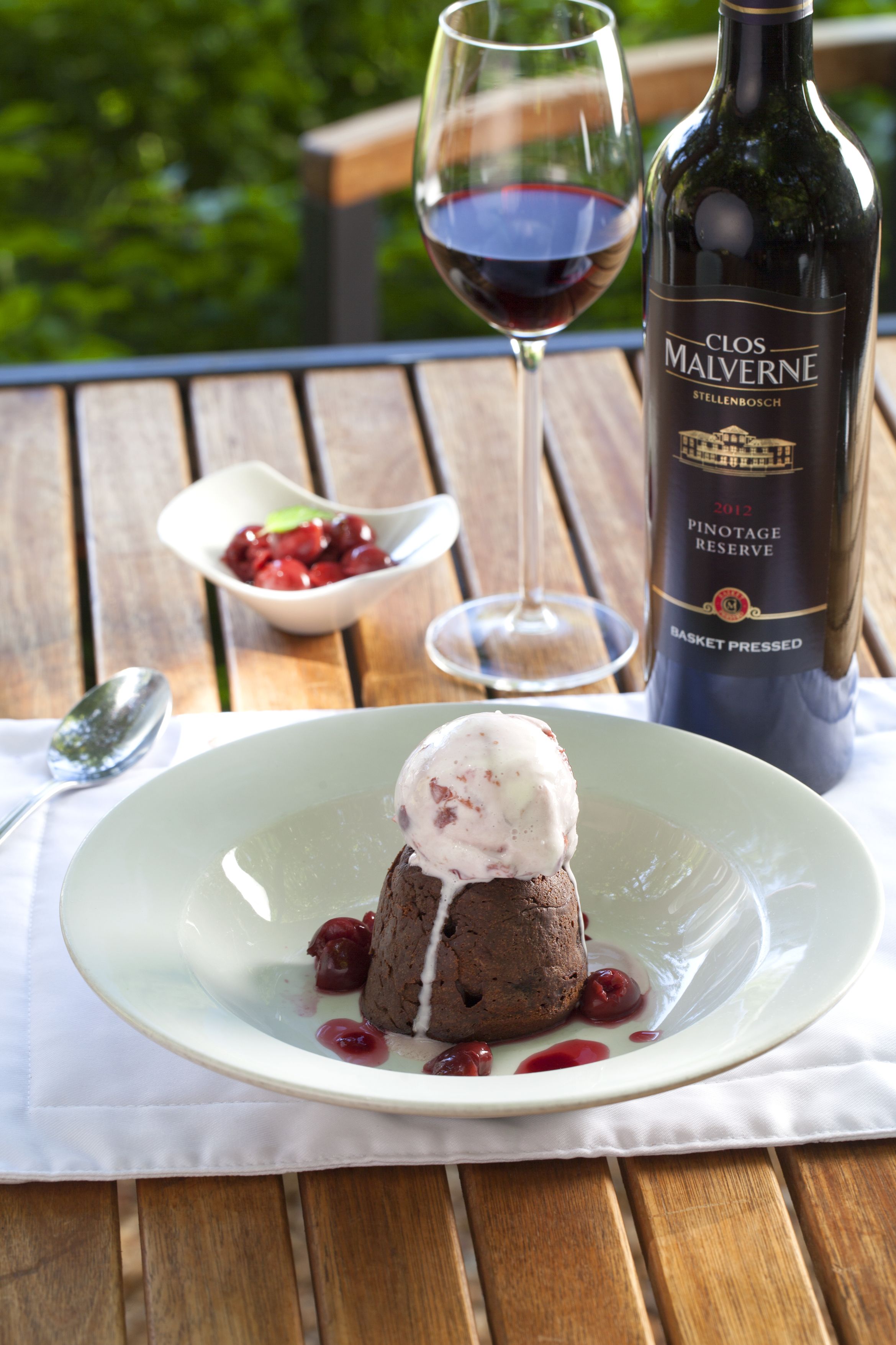 A dessert plate with a chocolate lava cake and berries, a wine glass, and a bottle of Clos Malverne wine on a wooden table.