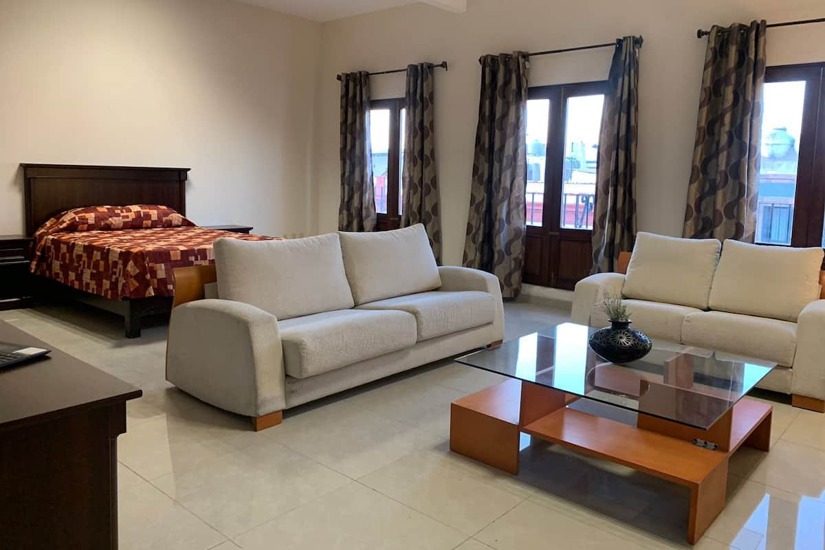 A modern living room with a sofa, a glass coffee table, and a bed with a patterned bedspread.