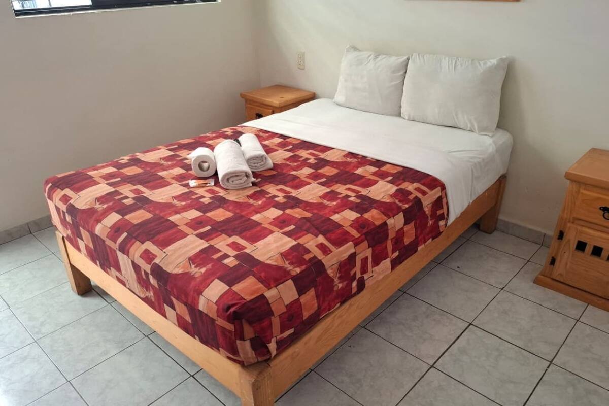 A single bed with a red and beige checkered bedspread, two pillows, and rolled towels on the bed.