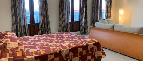 A bedroom with a large bed, a wooden dresser, and a view of the sea through the windows.