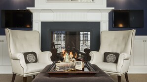 Un salon avec une cheminée, deux fauteuils, une table basse avec des livres et une bouteille, et une œuvre d’art encadrée au-dessus de la cheminée.