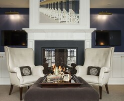 A living room with a fireplace, two armchairs, a coffee table with books and a bottle, and a framed artwork above the fireplace.