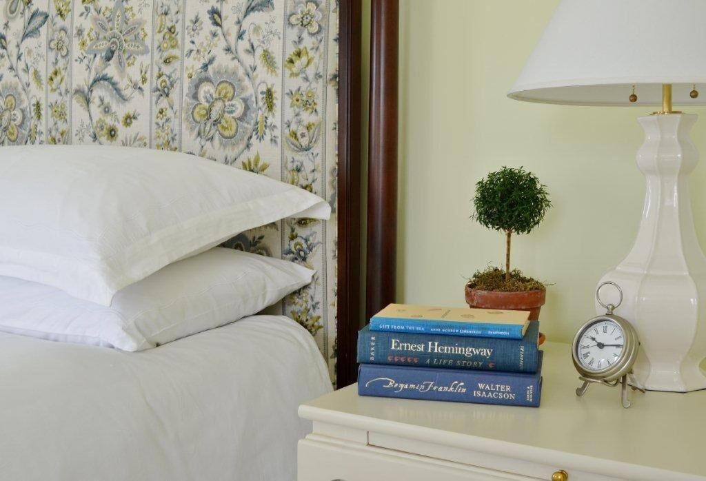 Una cama bien hecha con ropa blanca, una mesita de noche con lámpara, una pequeña planta en maceta y una pila de libros.