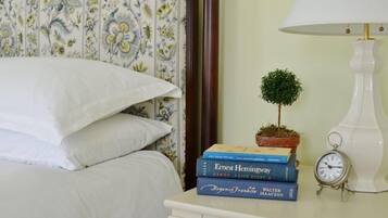 Una cama bien hecha con ropa blanca, una mesita de noche con lámpara, una pequeña planta en maceta y una pila de libros.
