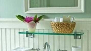 A bathroom with a white sink, two faucets, a mirror above the sink, a glass shelf with a plant and a basket of toiletries.