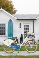 A bicycle with a basket and a surfboard leaning against a house.