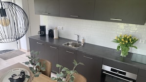 A modern kitchen with grey cabinets, a stainless steel sink, and an oven. There is a dining area with a wooden table and chairs, and a vase with yellow flowers on the counter.