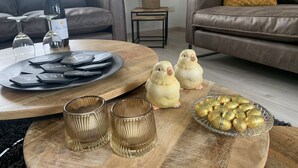 A cozy living room with a wooden coffee table, two decorative bird figurines, a tray with playing cards, a bottle of wine, and a bowl of chocolates.