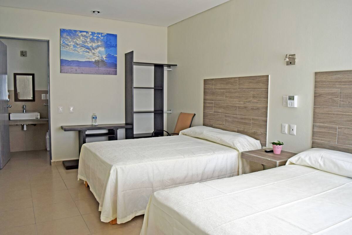Ein Hotelzimmer mit zwei Betten, einem Schreibtisch, einem Stuhl, einem kleinen Tisch mit Pflanze und einem im Hintergrund sichtbaren Badezimmer.