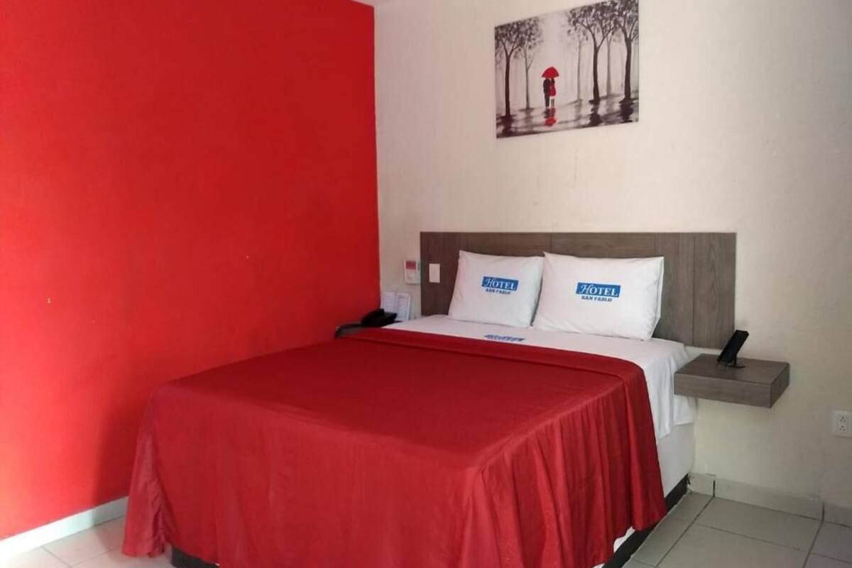 Ein Hotelzimmer mit roter Wand, einem Bett mit roter Bettdecke, zwei Kissen mit dem Hotelnamen, einem Nachttisch und einem Gemälde einer Person mit einem Regenschirm.