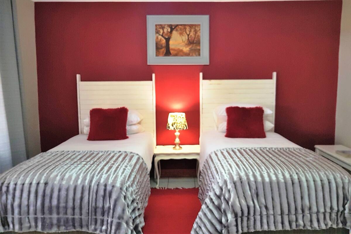 A hotel room with two beds, red walls, a painting of a tree, and a nightstand with a lamp.