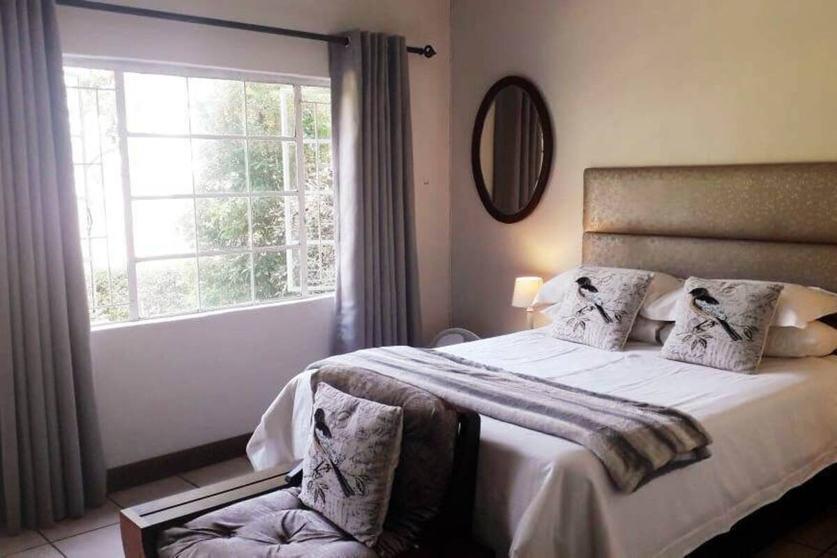A bedroom with a large bed, a chair, a window with curtains, and a nightstand with a lamp.