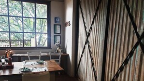 A dining area with a wooden table set for two, a window with a view of trees, and a corrugated metal wall. - Misty Mountains (Studholme)