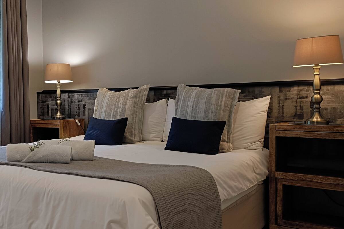 A hotel room with a large bed, two bedside tables with lamps, a view through sheer curtains, and a wooden headboard.