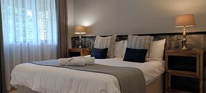 Habitación de hotel con una cama grande, dos mesitas de noche con lámparas, vistas a través de cortinas translúcidas y un cabecero de madera.