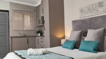 Une chambre à coucher comprenant un lit, une table de chevet, une fenêtre avec des stores, une porte et un petit coin cuisine.