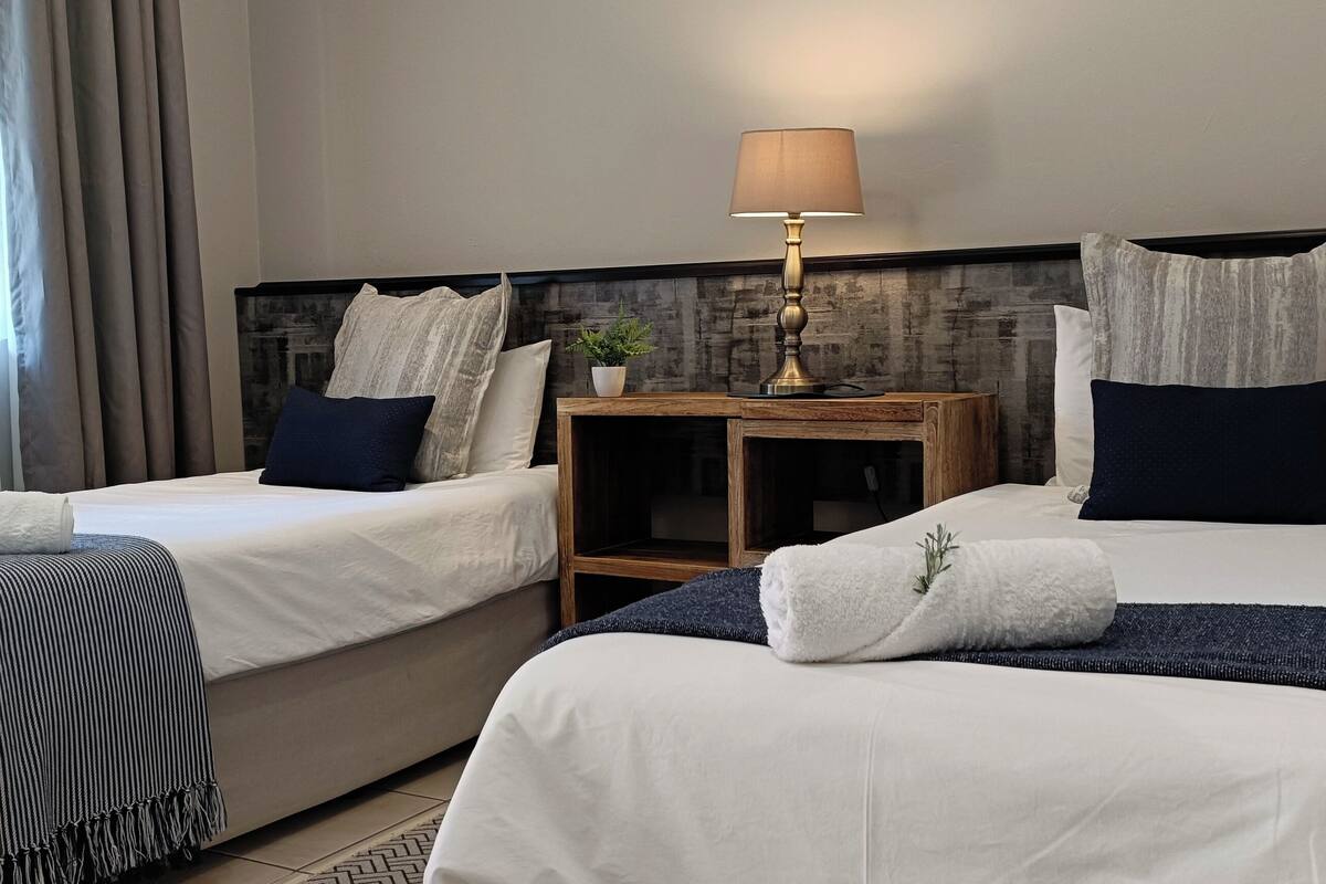 A hotel room with two beds, a nightstand with a lamp, and a view of the outdoors through the window.