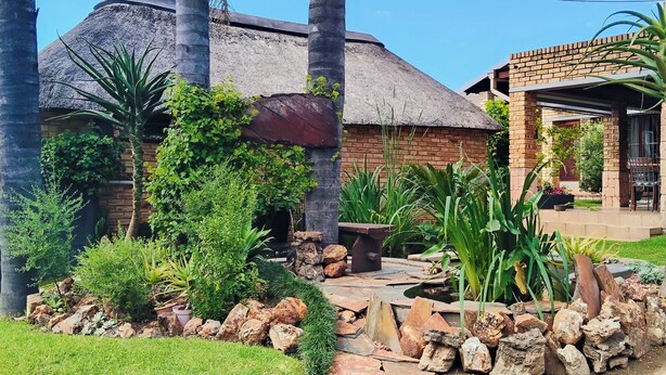 Una casa con techo de paja, rodeada por un jardín con rocas, plantas y un pequeño estanque.