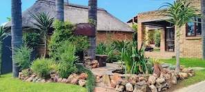 Una casa con techo de paja, rodeada por un jardín con rocas, plantas y un pequeño estanque.