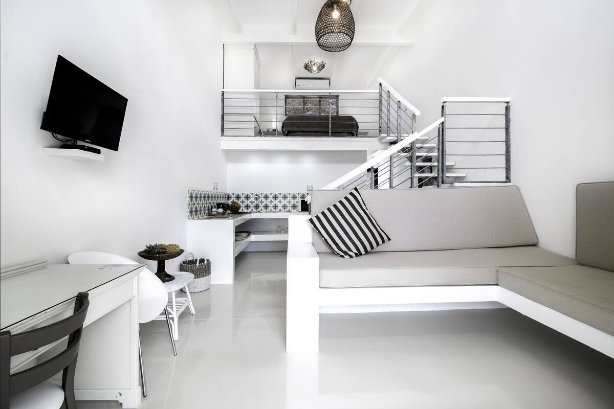 Un espace de vie moderne avec un téléviseur à écran plat fixé au mur, un coin repas avec une table et des chaises blanches, un canapé agrémenté d’un coussin à rayures et un escalier menant à un niveau supérieur.