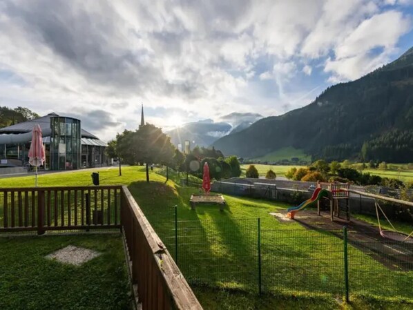 A playground with slides and a fenced area, surrounded by a grassy field and a mountain in the background.