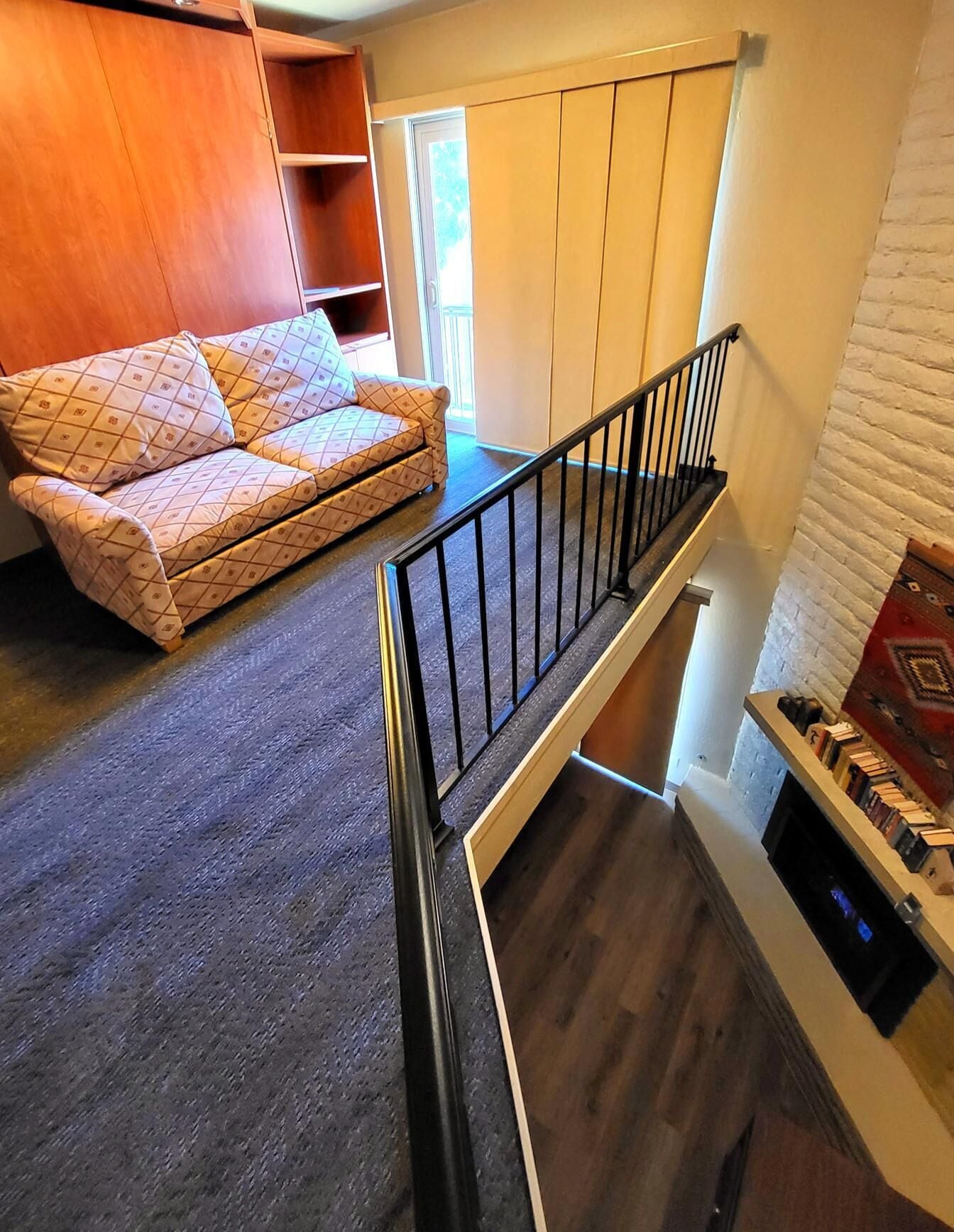 A loft-style room with a high ceiling, a built-in wooden wardrobe, a sofa with patterned cushions, and a staircase with a black railing.