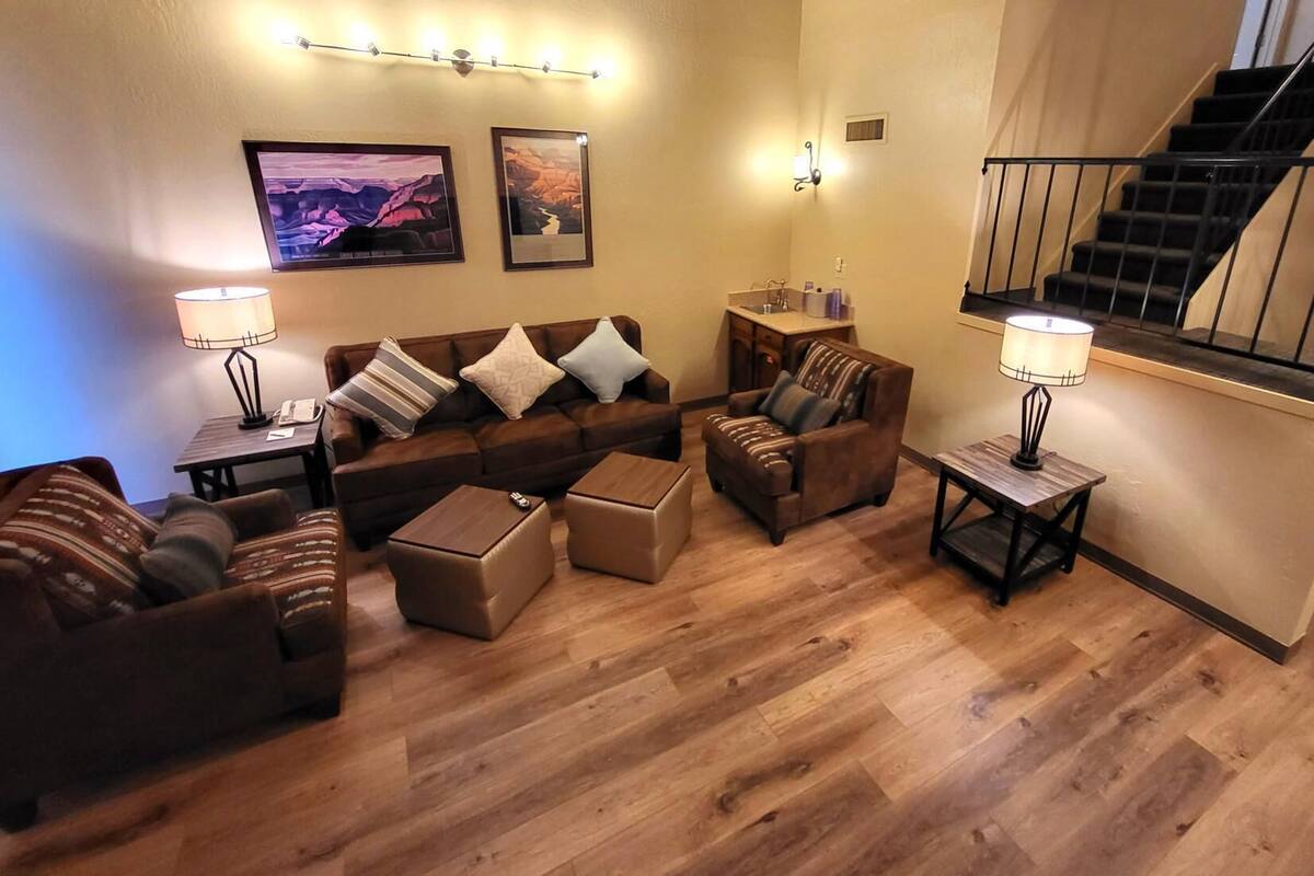 A cozy living room with a sofa set, ottomans, and a coffee table. There is a staircase to the right and a wall-mounted light fixture above the sofa.