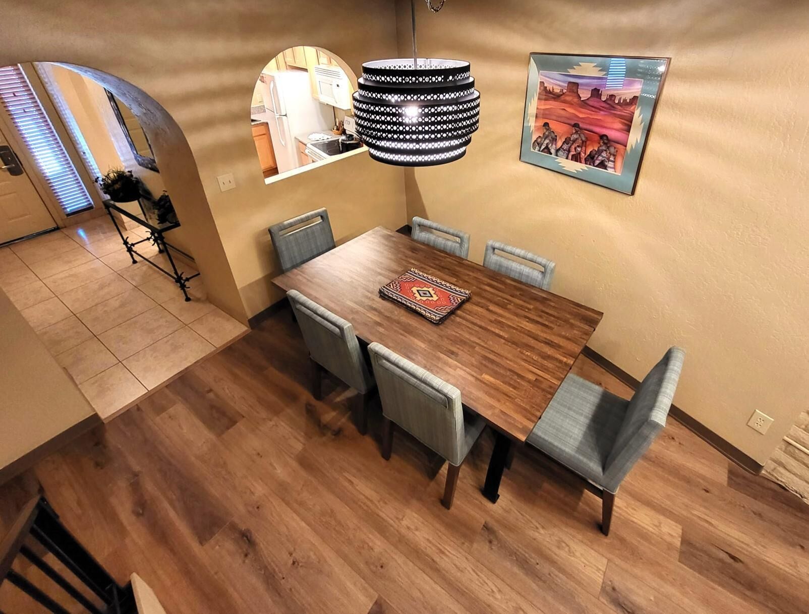 Un comedor con una mesa de madera, seis sillas, un tapete estampado, una lámpara colgante y un cuadro en la pared.