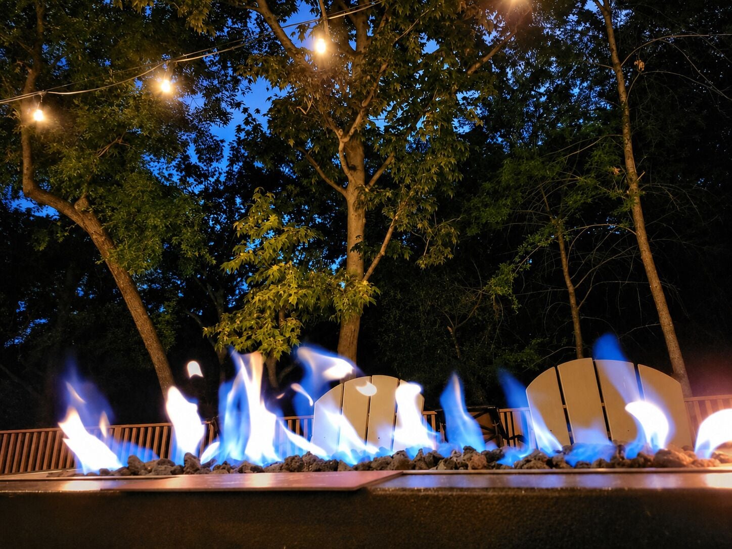A nighttime outdoor fire feature with flames, surrounded by trees and string lights.