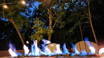 A nighttime outdoor fire feature with flames, surrounded by trees and string lights.