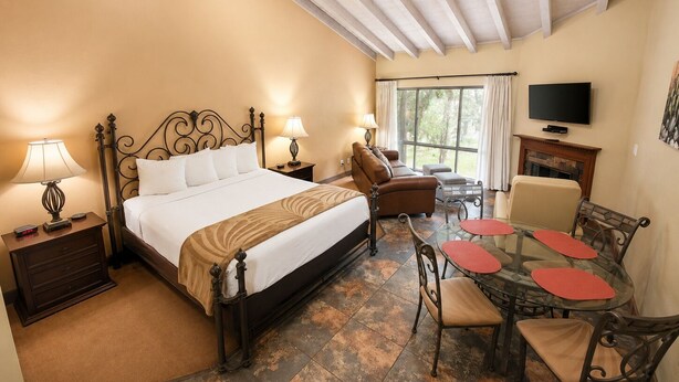 A hotel room with a bed, bedside lamps, a sofa, a dining table with chairs, a television, and a fireplace. - Villas At Poco Diablo (Sedona)