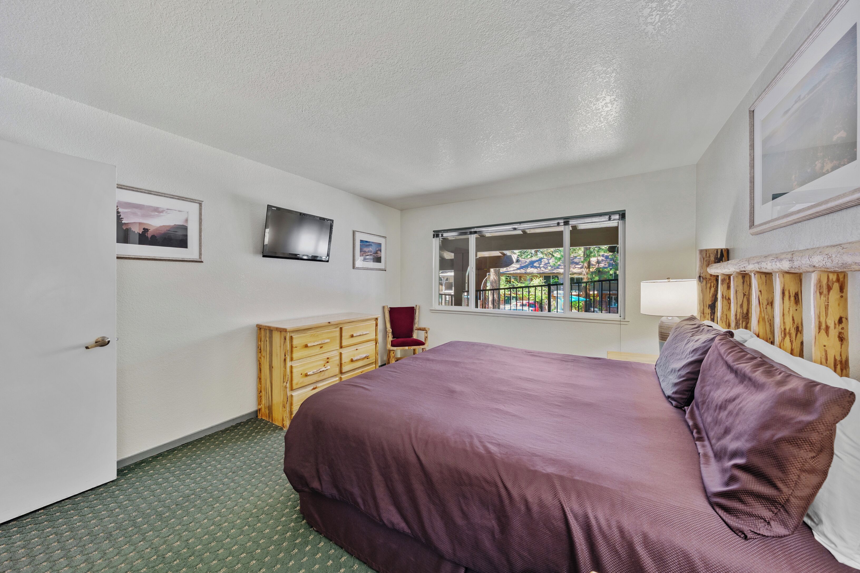 A bedroom with a large bed, a wooden dresser, a chair, a TV, and a window with a view of greenery.
