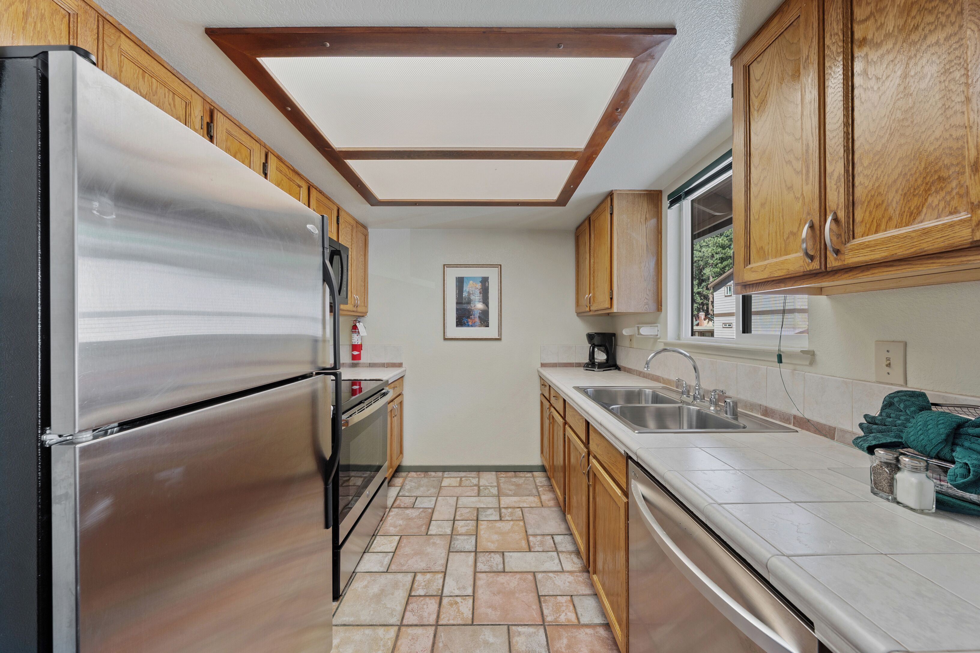 A kitchen with a stainless steel refrigerator, wooden cabinets, a tiled floor, a window, and a sink.