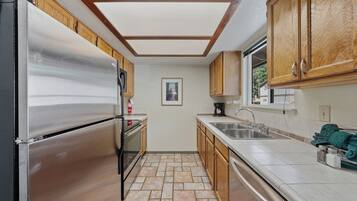 A kitchen with a stainless steel refrigerator, wooden cabinets, a tiled floor, a window, and a sink.